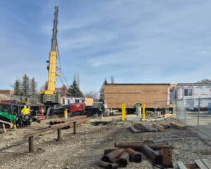 Land surveyors on an active construction site coordinating work around a crane, steel piles, and heavy machinery during site preparation.